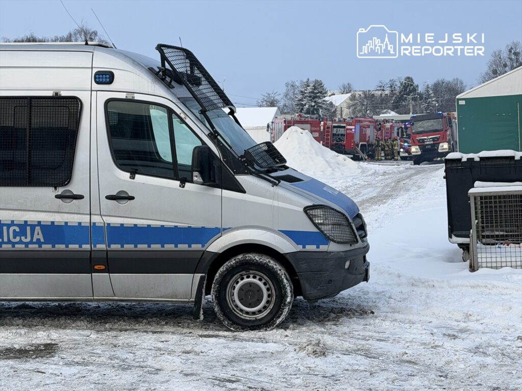 Policyjny radiowóz stoi na zaśnieżonym terenie obok wozów strażackich i grupy strażaków w akcji.