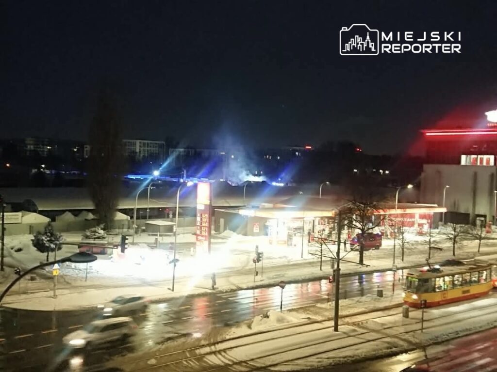 Świecące neony stacji benzynowej w zimowej scenerii nocnej, z ośnieżonymi ulicami i przejeżdżającym tramwajem.