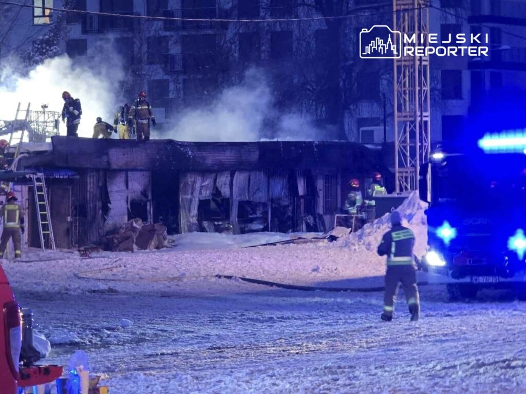 Grupa strażaków gasi pożar w zniszczonym budynku otoczonym śniegiem, w tle widać niebieskie światła wozu strażackiego.