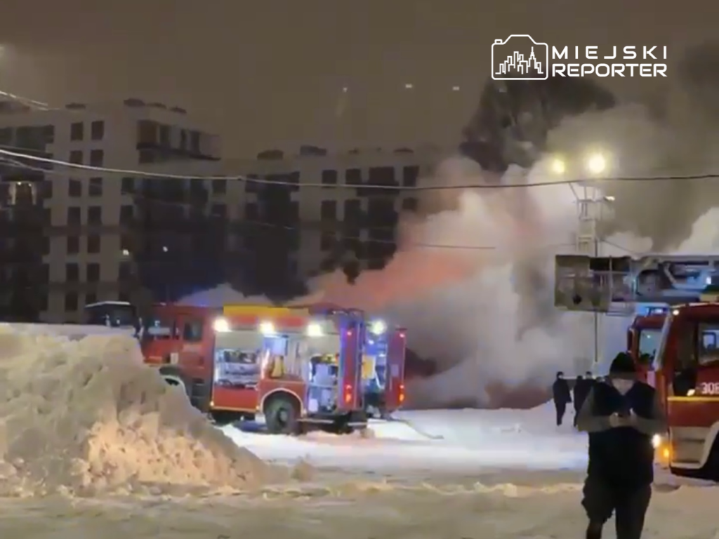 Strażacy z wozów gaśniczych walczą z pożarem w zaśnieżonym terenie, w tle nowoczesne budynki mieszkalne.