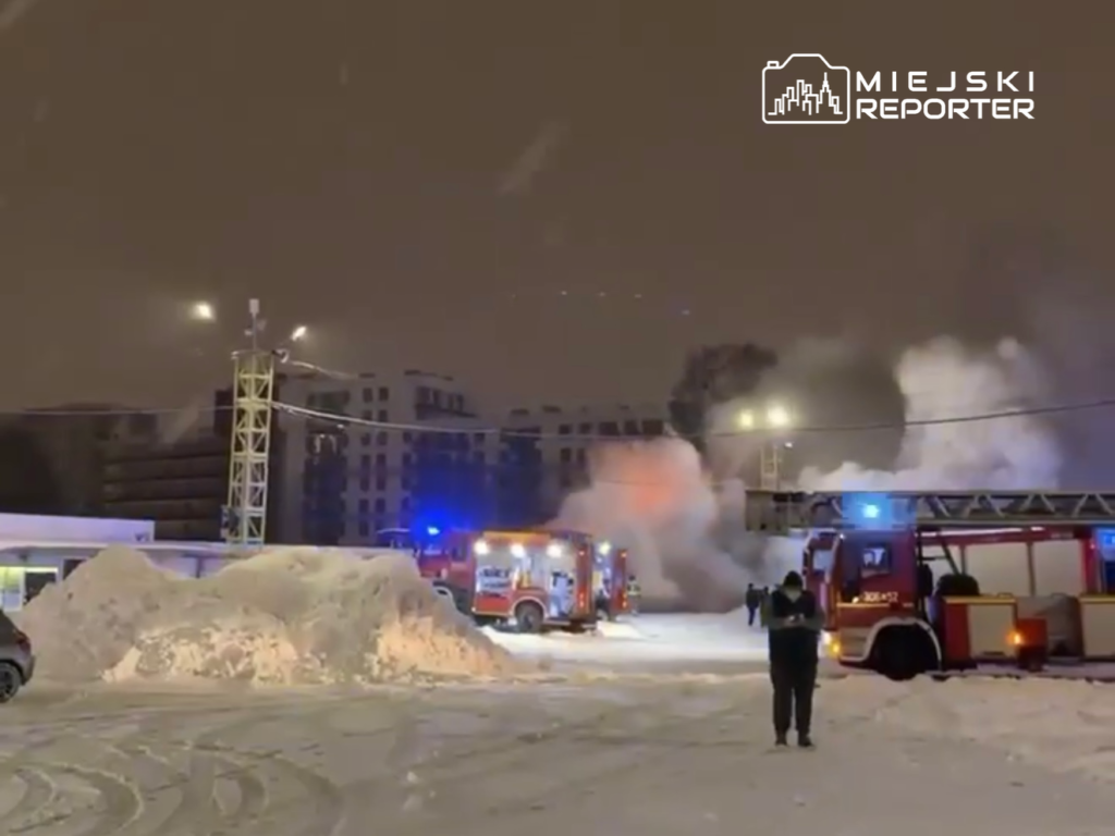 Strażacy z wozami bojowymi gaszą pożar w zaśnieżonym terenie miejskim w nocy. W tle widoczne są budynki.