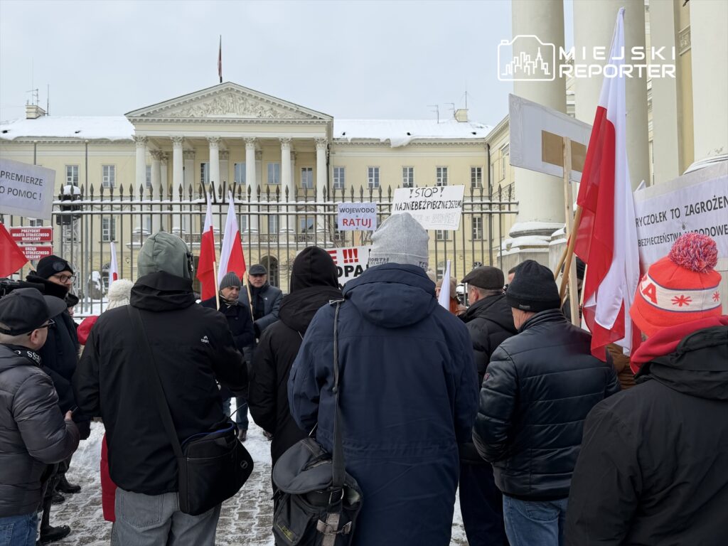 Grupa protestujących z flagami narodowymi i transparentami stoi przed budynkiem rządowym w zimowej scenerii.