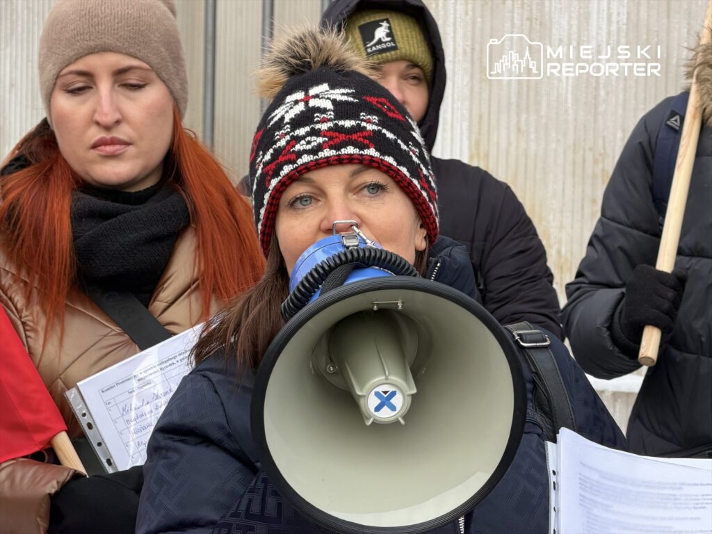 Kobieta w czapce z pomponem przemawia przez megafon, stojąc w grupie osób trzymających transparenty.