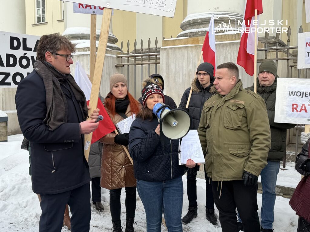 Grupa protestujących, w tym kobieta z megafonem, stoi na śniegu przed budynkiem z transparentami i flagami.