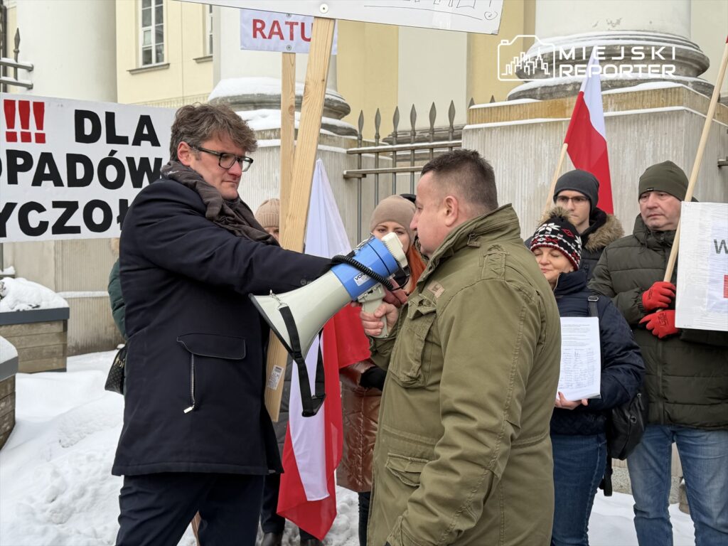 Mężczyzna w garniturze trzymający megafon przemawia do zgromadzonych protestujących w zimowej scenerii, obok transparenty.