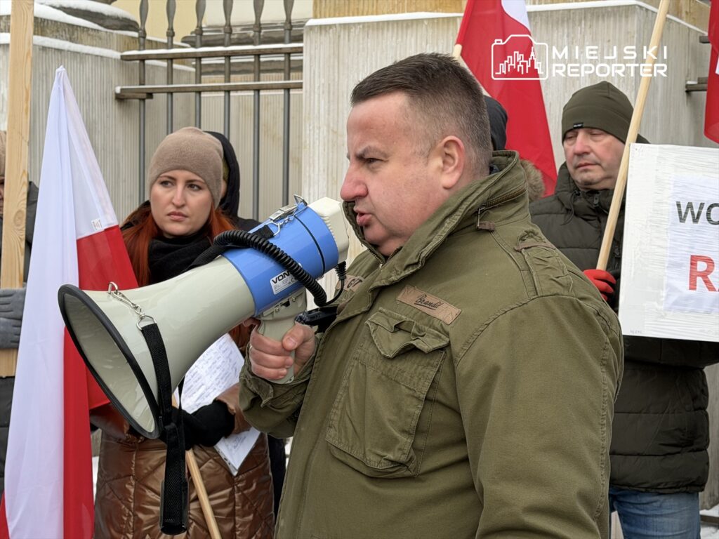 Mężczyzna w kurtce wojskowej przemawia przez megafon, a w tle kobieta trzyma flagę Polski. Wszyscy uczestnicy protestu są ubrani w ciepłe kurtki.