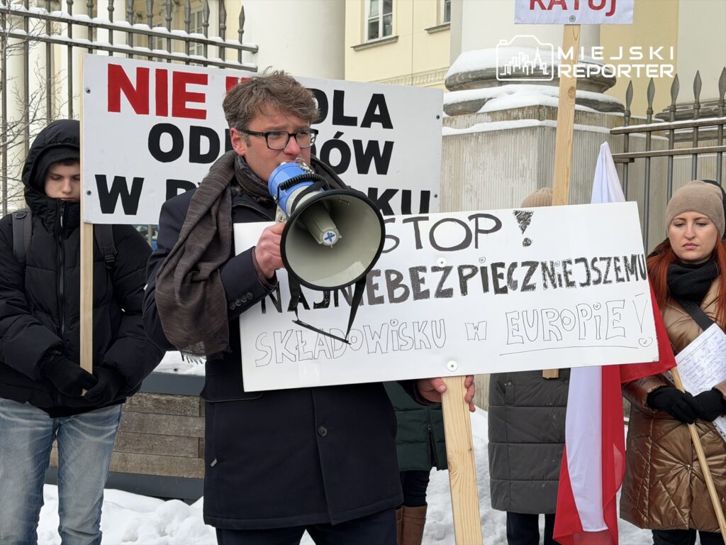 Mężczyzna w garniturze przemawia przez megafon podczas protestu, a w tle widoczne transparenty z hasłami.