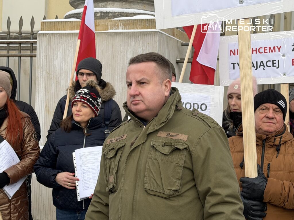 Grupa protestujących z transparentami i flagami narodowymi stoi na zewnątrz, w tle widoczne budynki.