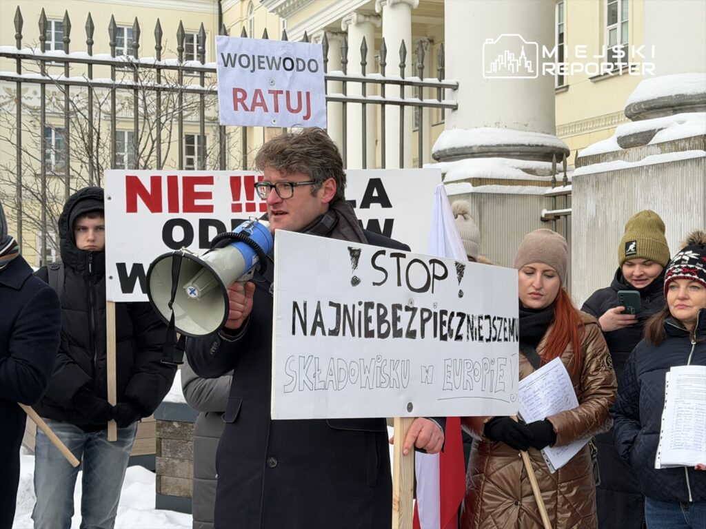 Mężczyzna w okularach trzymający megafon przemawia do grupy protestujących, trzymających transparenty w zimowym otoczeniu.
