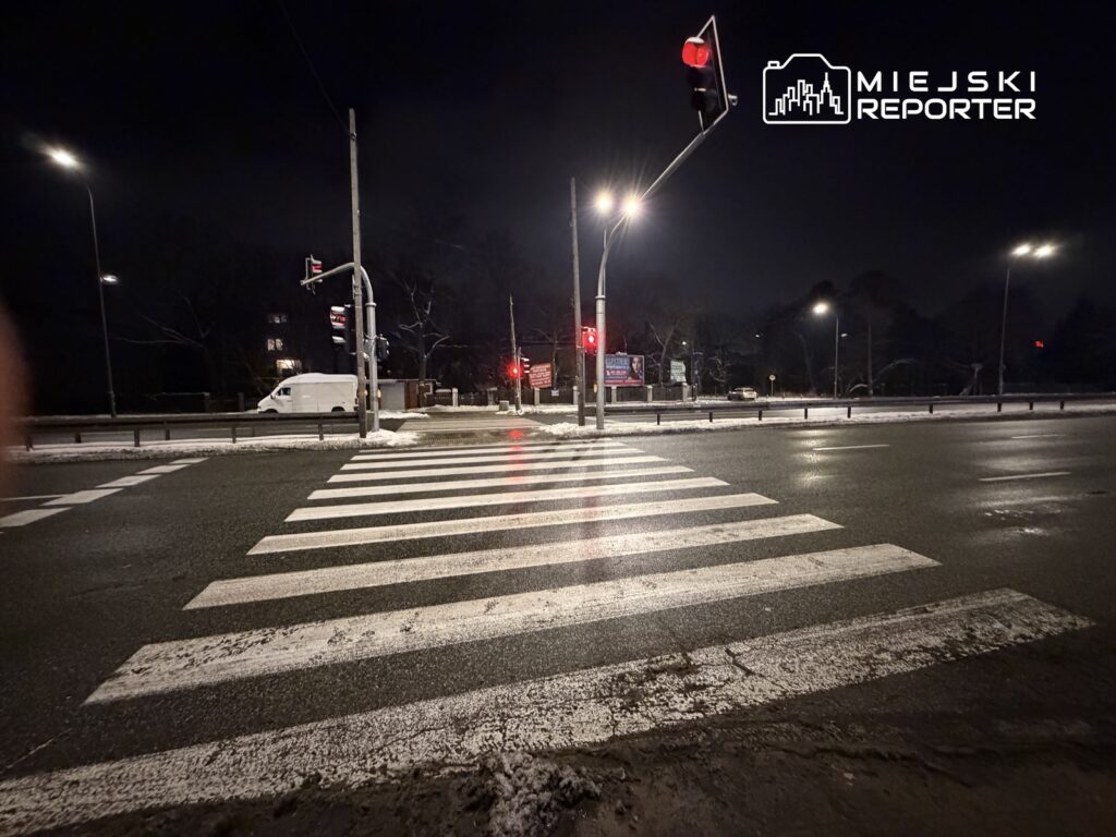 Zebrażdżona przejście dla pieszych oświetlona latarniami w nocnej scenerii, z zapalonym czerwonym światłem sygnalizacji.