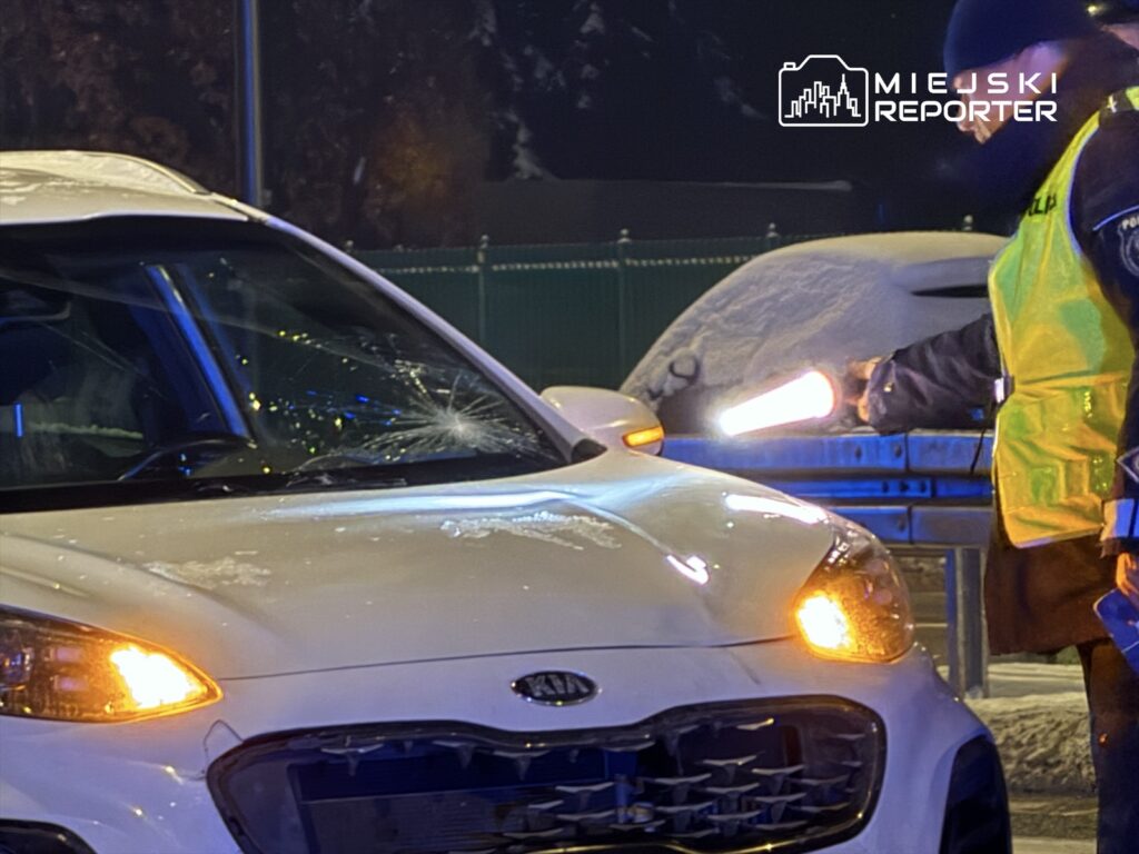 Policjant w odblaskowej kamizelce kontroluje uszkodzony samochód osobowy w zimowej scenerii.