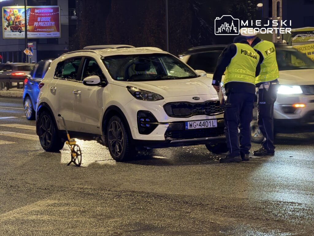 Policjanci w odblaskowych kamizelkach badają uszkodzony samochód osobowy na ruchliwej ulicy nocą.