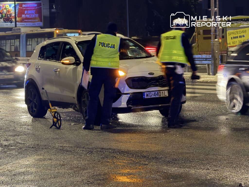 Policjanci w odblaskowych kamizelkach badają uszkodzony samochód na mokrej jezdni, używając narzędzia pomiarowego.