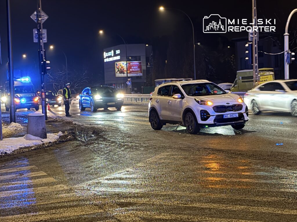 Policjant kierujący ruchem drogowym na skrzyżowaniu, obok pojazdów z włączonymi światłami.