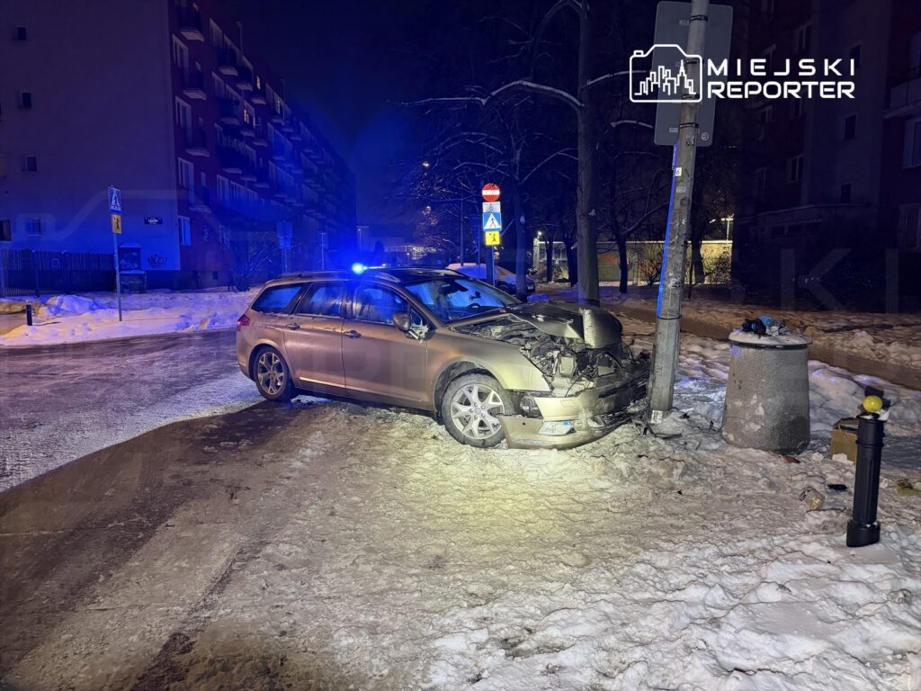 Uszkodzony samochód osobowy stoi na skrzyżowaniu w nocy, obok przystanku z niebieskimi światłami policyjnymi.