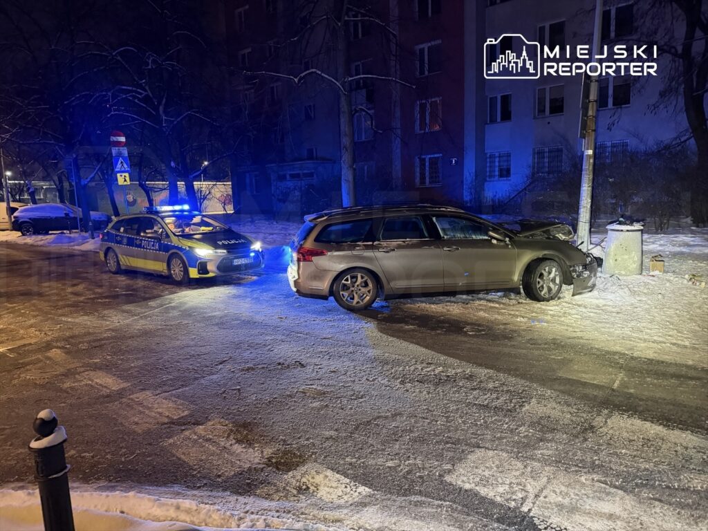 Policyjny radiowóz z włączonymi sygnałami świetlnymi stoi obok uszkodzonego samochodu, który uderzył w latarnię, na ośnieżonej ulicy w nocy.