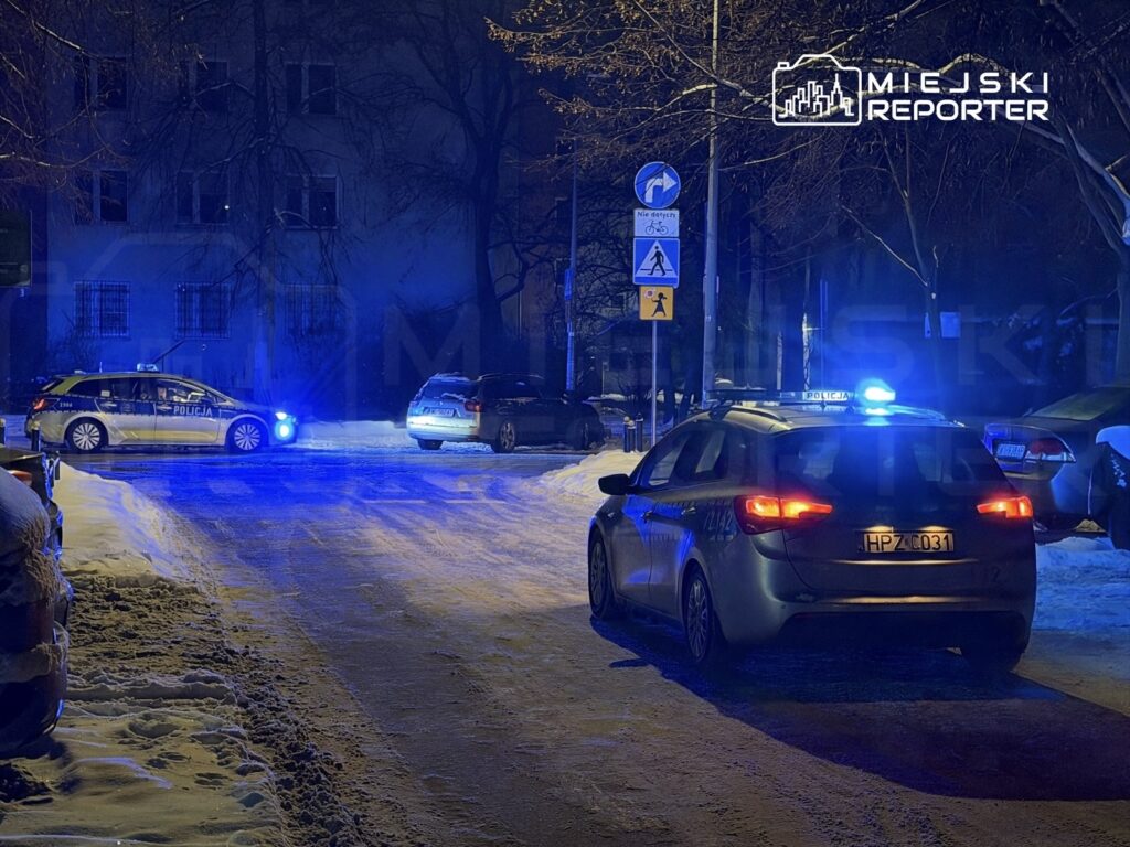 Policyjny samochód z włączonymi sygnałami świetlnymi stoi na ośnieżonej ulicy, obok drugiego pojazdu.