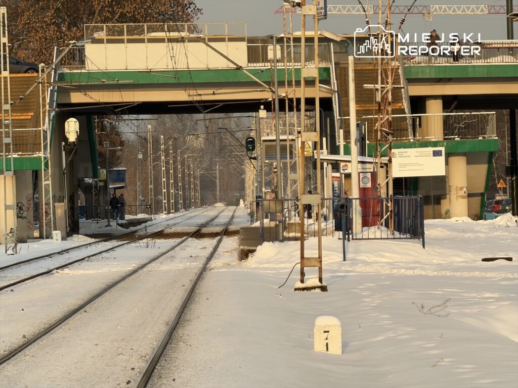 Zimowy krajobraz stacji kolejowej z torami pokrytymi śniegiem, semaforem i pasażerami oczekującymi na pociąg.