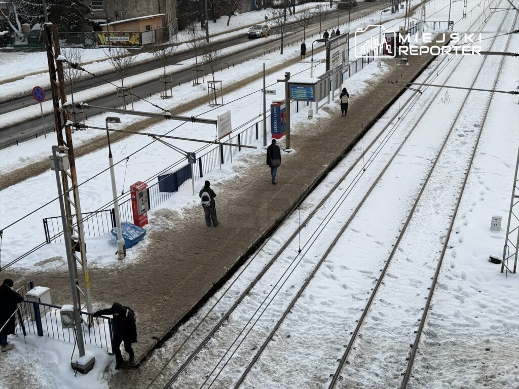 Pasażerowie czekają na przystanku tramwajowym na zaśnieżonym terenie, obok torów tramwajowych.