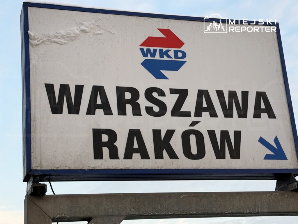 Tablica informacyjna z napisem "WARSZAWA RAKÓW" oraz symbolami WKD, umieszczona na metalowej konstrukcji.