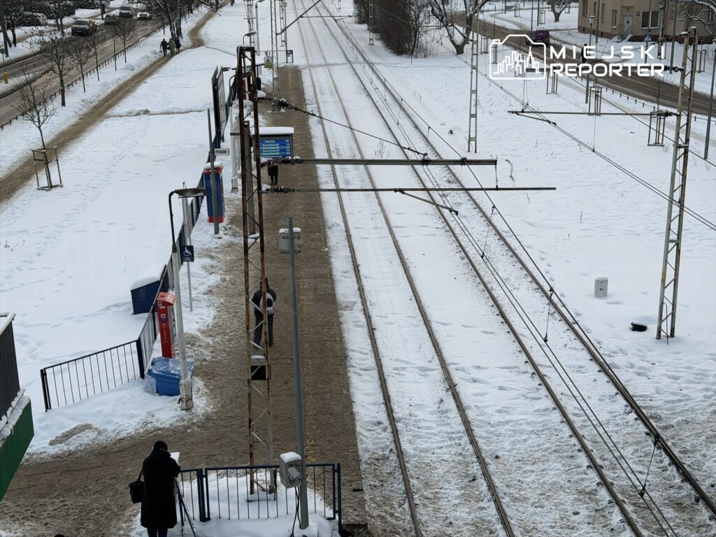 Osoby czekające na peronie w zimowej scenerii, otoczone śniegiem i torami kolejowymi.