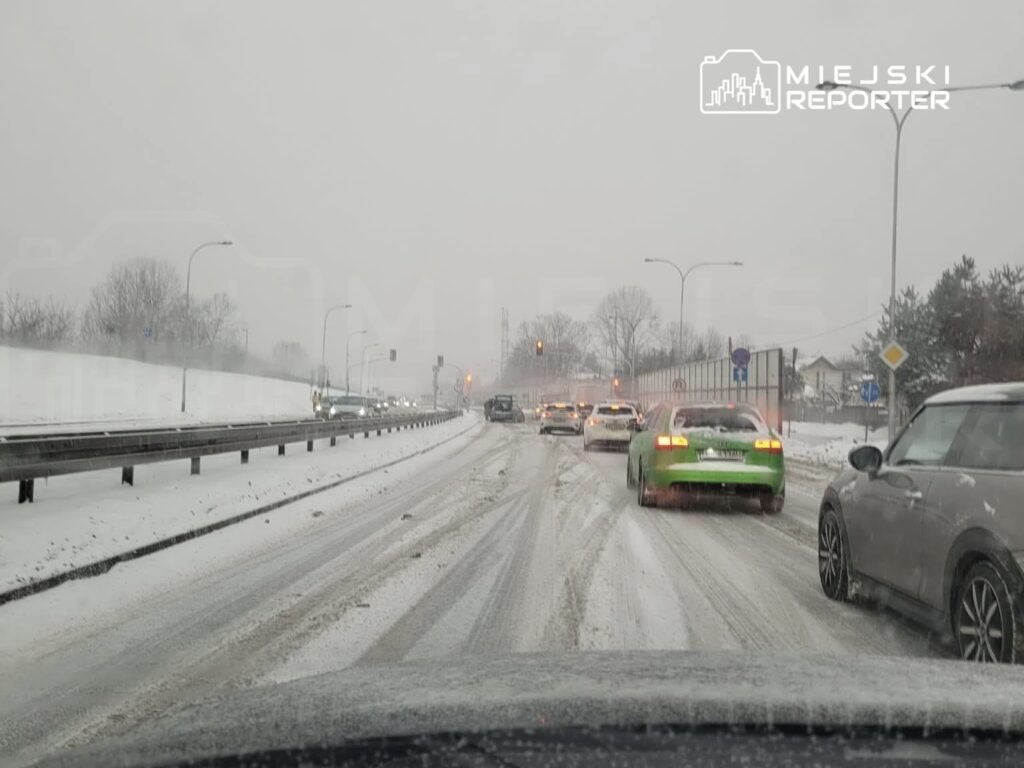 Ruch samochodowy na zaśnieżonej drodze, z widocznymi światłami sygnalizacyjnymi i drzewami w tle.