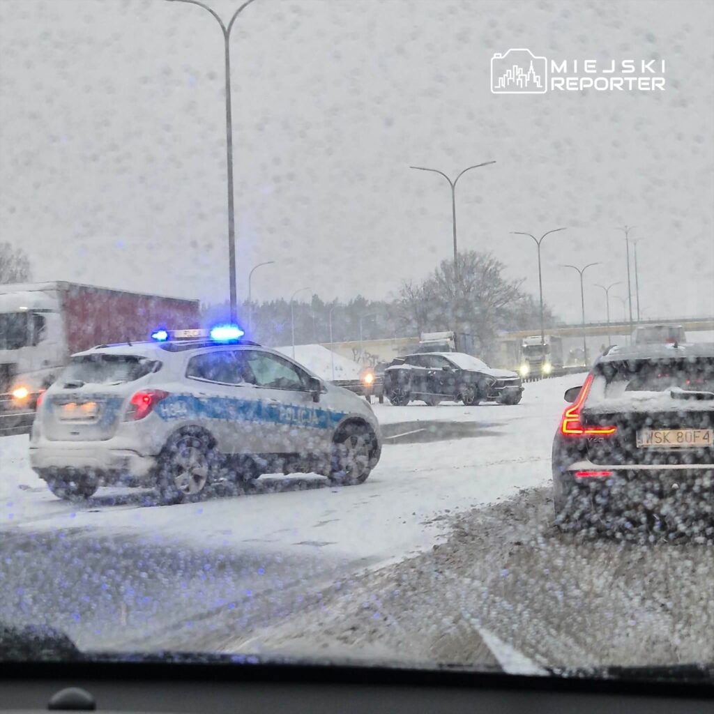 Policyjny radiowóz z włączonymi sygnałami świetlnymi stoi na zaśnieżonej drodze, obok widoczne inne pojazdy.