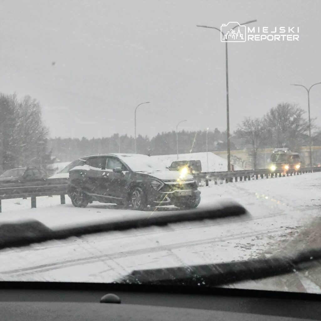 Samochód utknął w śniegu na drodze, wokół panują warunki zimowe z opadami śniegu i ograniczoną widocznością.