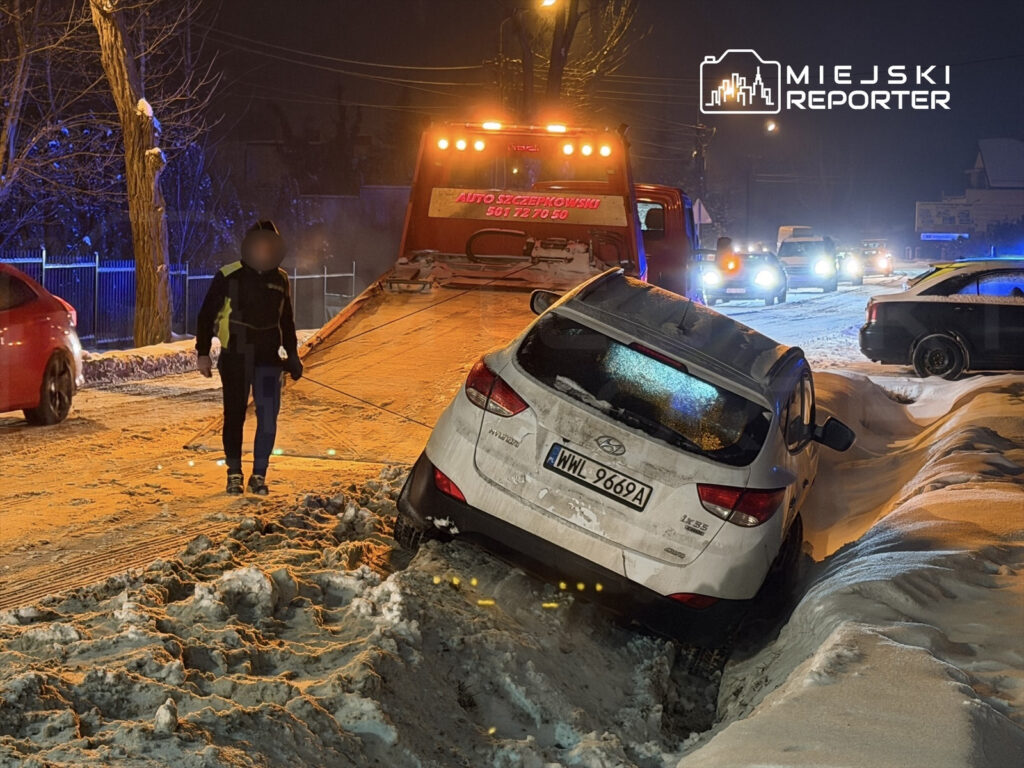 Mężczyzna w odzieży roboczej kieruje holowaniem samochodu osobowego utknęłego w śniegu na oświetlonej ulicy.