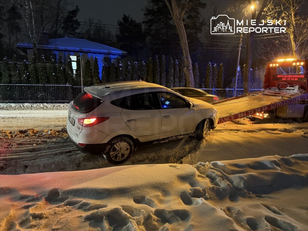 Biały samochód osobowy utknął w śniegu obok drogi, podczas gdy laweta podjeżdża, aby go odholować. W tle widoczny dom i oświetlenie uliczne.