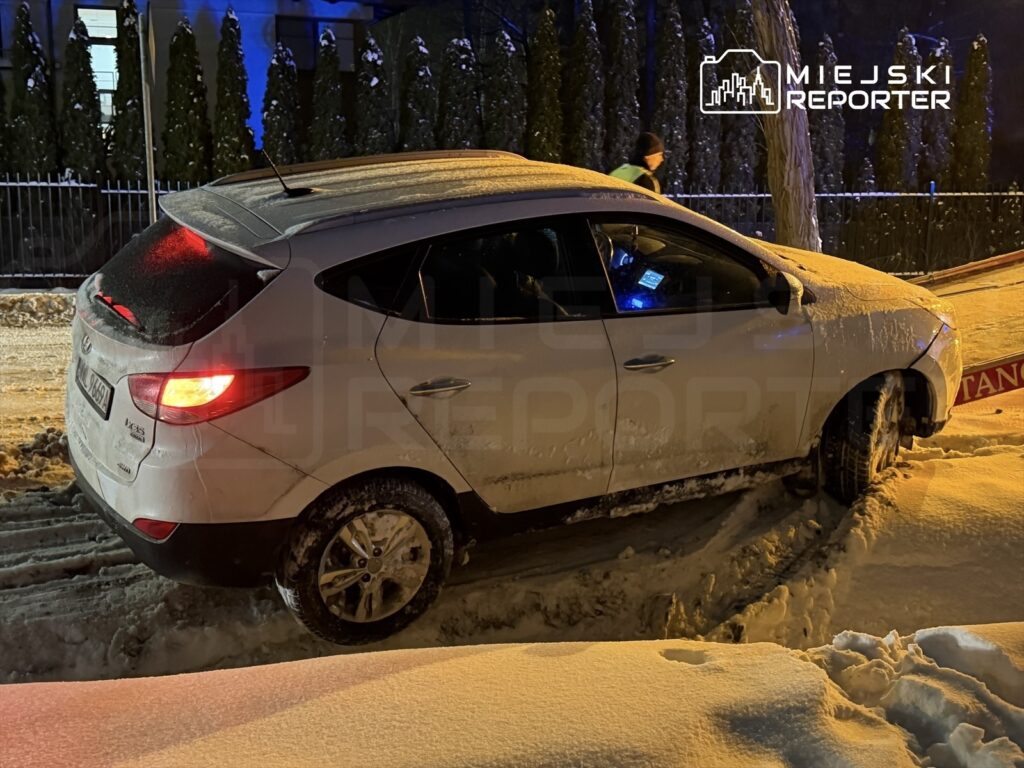 Samochód osobowy utknął w śniegu na zaśnieżonej ulicy, obok przechodzi osoba w kurtce.