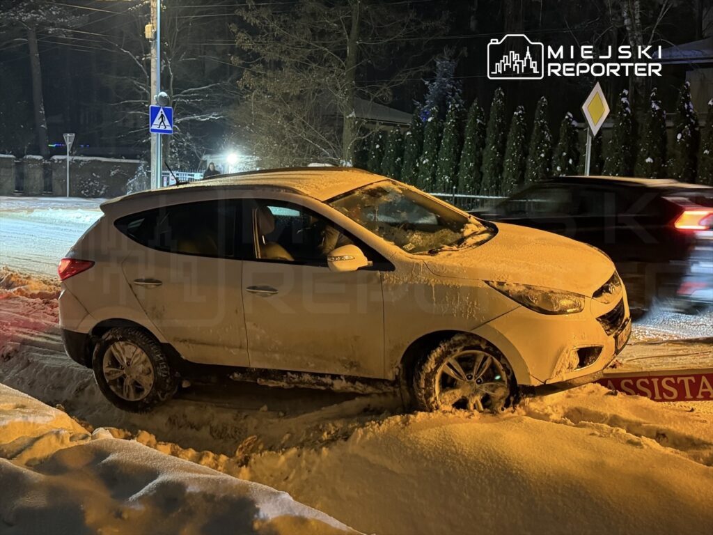 Biały samochód osobowy utknął w śniegu na poboczu, obok znaku drogowego i oświetlonej ulicy.