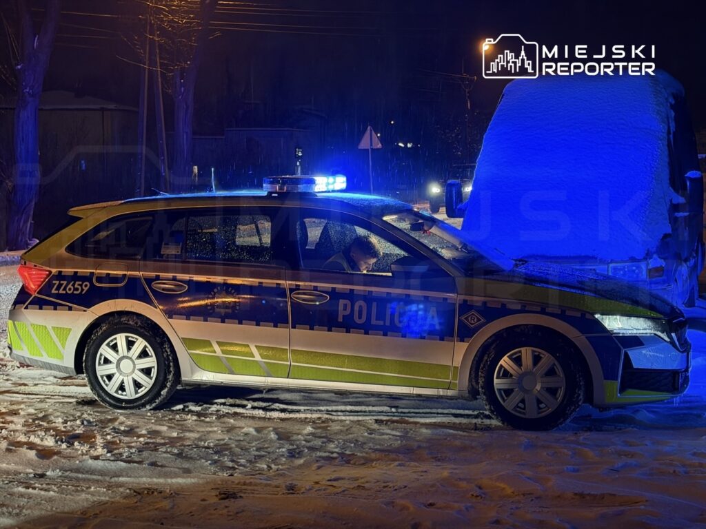 Policyjny radiowóz z włączonymi sygnalizatorami świetlnymi stoi na zaśnieżonej drodze w nocy.