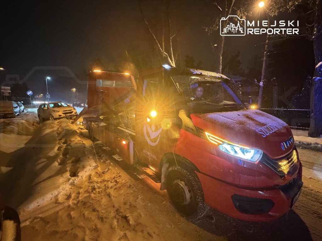 Czerwony holownik z włączonymi światłami stoi na zaśnieżonej ulicy, obok zaparkowanych samochodów.