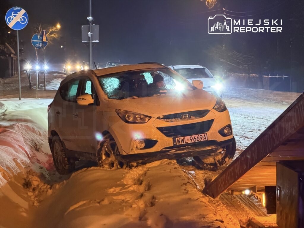 Osobowy samochód naśnieżonym poboczu, z jedną osią uniesioną, w tle oświetlona ulica i znaki drogowe.