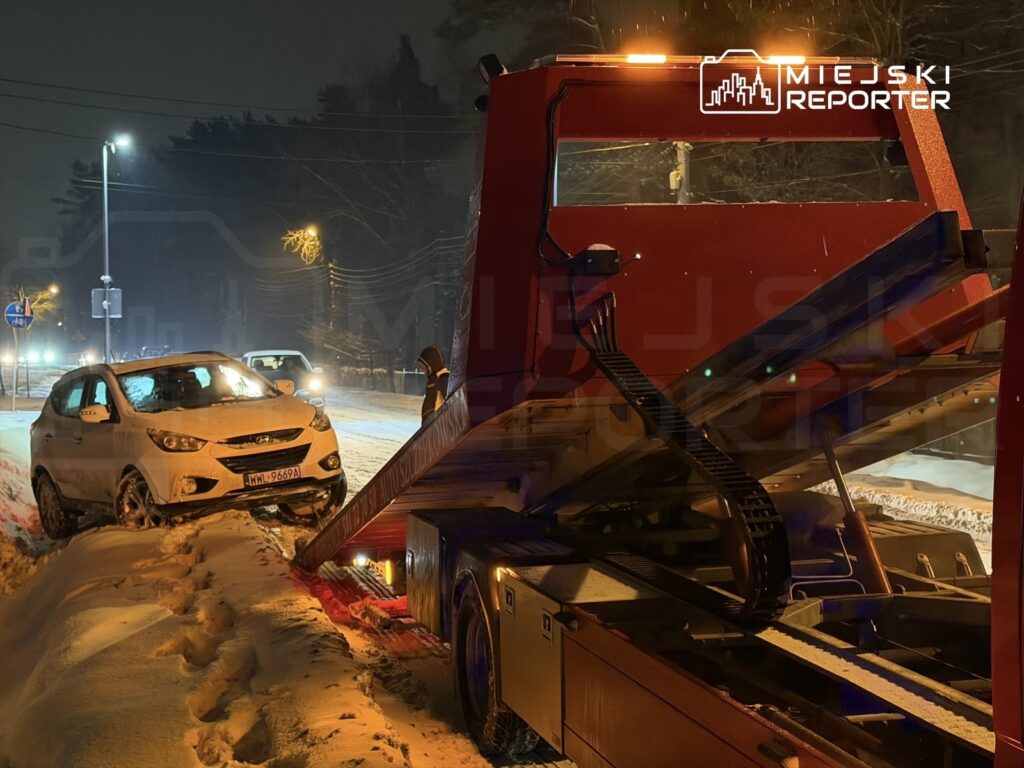 Samochód osobowy utknął w śniegu, podczas gdy laweta podjeżdża, aby go odholować w zimowej scenerii nocą.
