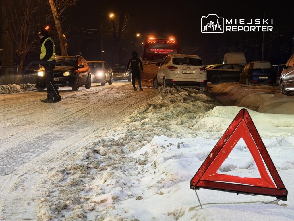 Grupa osób w odblaskowych kamizelkach kieruje ruchem na zaśnieżonej drodze w nocy, obok stoi trójkąt ostrzegawczy.