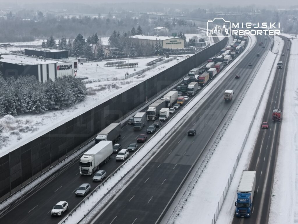 Ruch samochodowy na zaśnieżonej drodze ekspresowej z zatorami, w tle budynki i drzewa pokryte śniegiem.