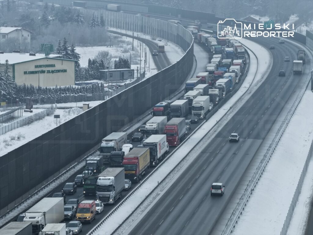 Kolejka ciężarówek i osobowych samochodów zatrzymana na zaśnieżonej drodze, otoczona wysokim murem.