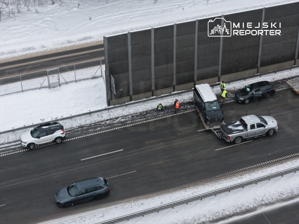 Na drodze pokrytej śniegiem, zespół ratunkowy pracuje przy uszkodzonym samochodzie, obok zaparkowanej ciężarówki.