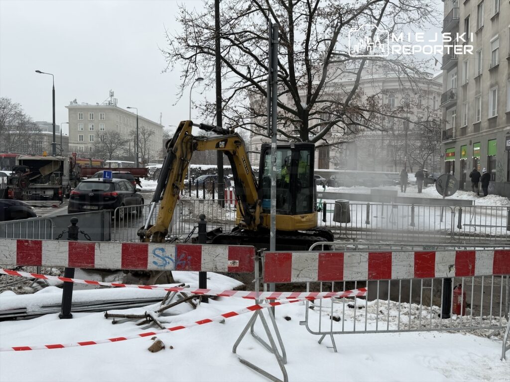 Maszyna budowlana kopie w śniegu na ulicy, otoczona barierkami i taśmą ostrzegawczą, w tle samochody i budynki.