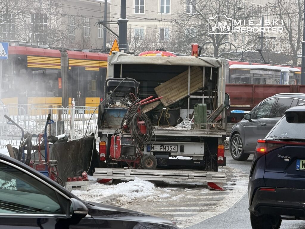 Pojazd roboczy z narzędziami stoi na zaśnieżonej ulicy, obok przejeżdża tramwaj.