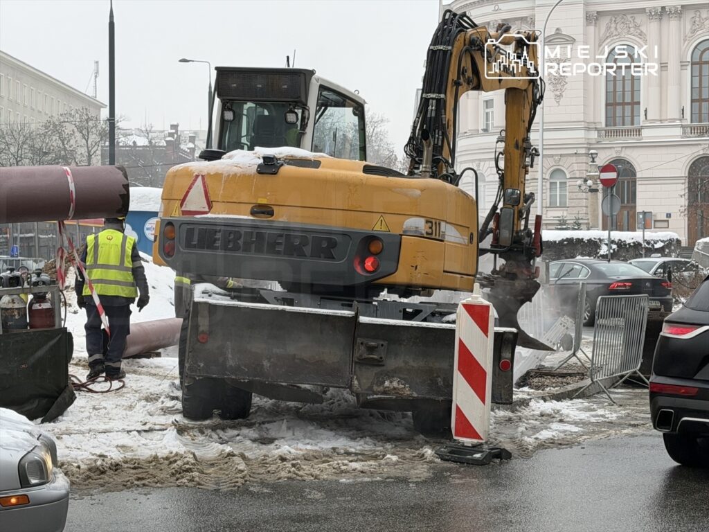 Robotnik w kamizelce odblaskowej obsługuje maszynę budowlaną na ośnieżonej ulicy.