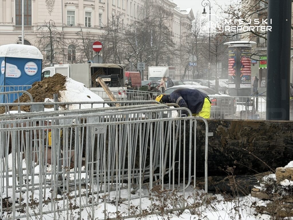 Robotnik w żółtej odzieży roboczej wykonuje prace przy wykopie w zaśnieżonym mieście, otoczony barierkami.