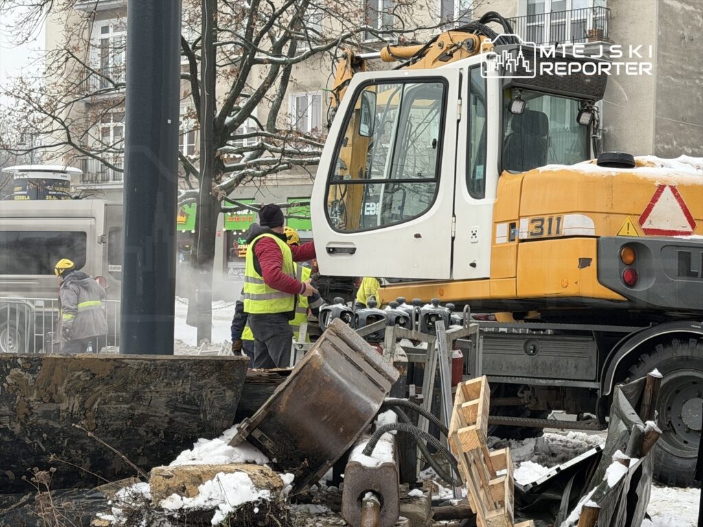 Grupa pracowników w odblaskowych kamizelkach prowadzi prace budowlane w zimowym otoczeniu, obok dużej maszyny budowlanej.