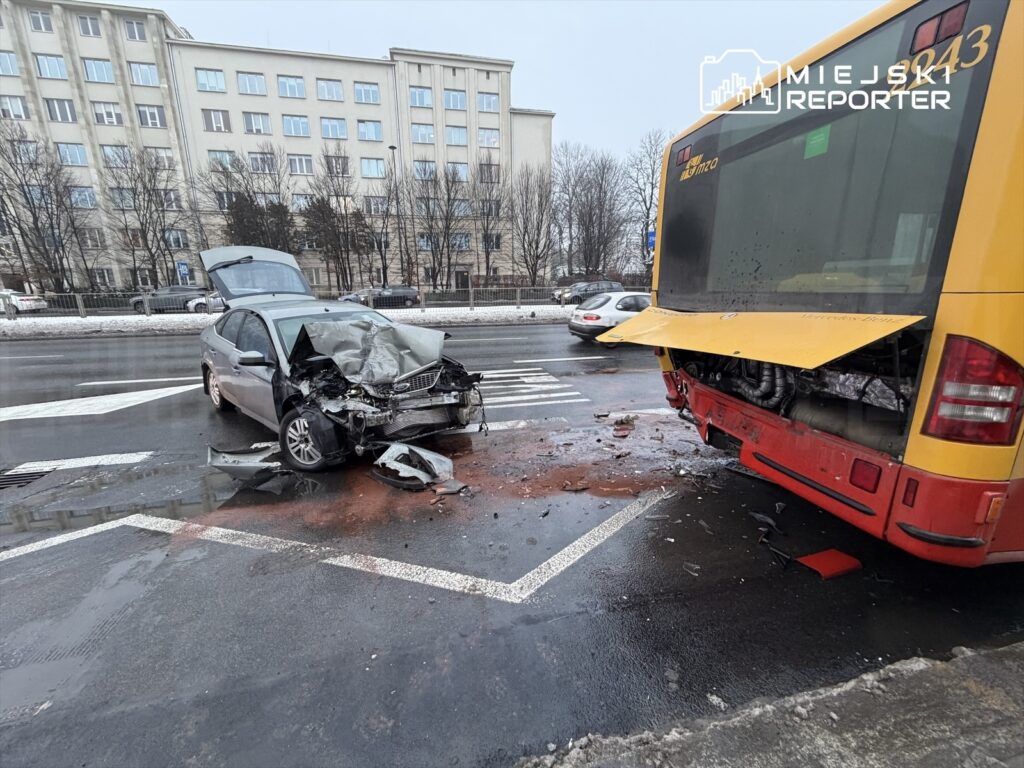 Zniszczony samochód osobowy z wgniecionym przodem stoi obok uszkodzonego autobusu na mokrej jezdni.