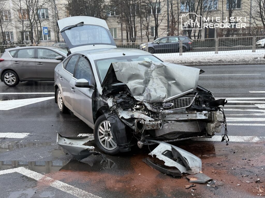 Uszkodzony srebrny samochód osobowy z odgiętym dachem stoi na pasach, obok widać fragmenty karoserii i plamę na jezdni.