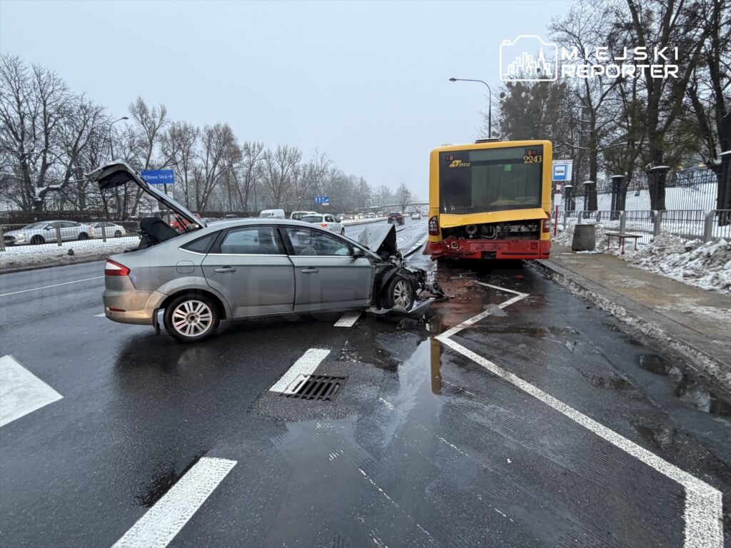 Zniszczony samochód osobowy z otwartą pokrywą bagażnika stoi na jezdni obok uszkodzonego autobusu w zimowej scenerii.