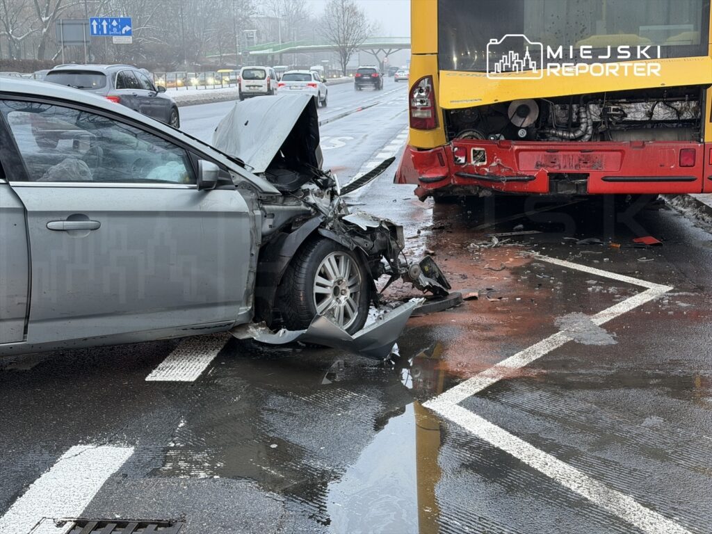 Zderzenie samochodu osobowego z autobusem na mokrej nawierzchni drogi, w tle ruchliwe ulice i znaki drogowe.