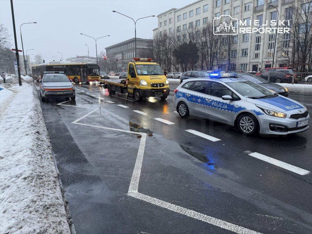 Policyjny radiowóz z włączonymi sygnałami świetlnymi oraz laweta zatrzymane na mokrej ulicy, obok przystanku autobusowego.